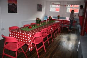 Two long tables with red folding chairs and red spot table cloth. two guests setting room up for event